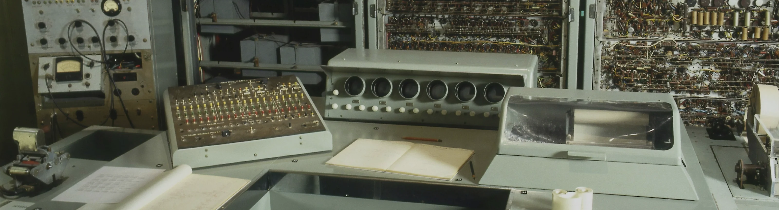 CSIRAC, a 1950s room-sized computer at Museums Victoria: the operator's console and desk with open notebooks, in front of grey cabinets of hand-wired racks. Photo by Museums Victoria on Unsplash